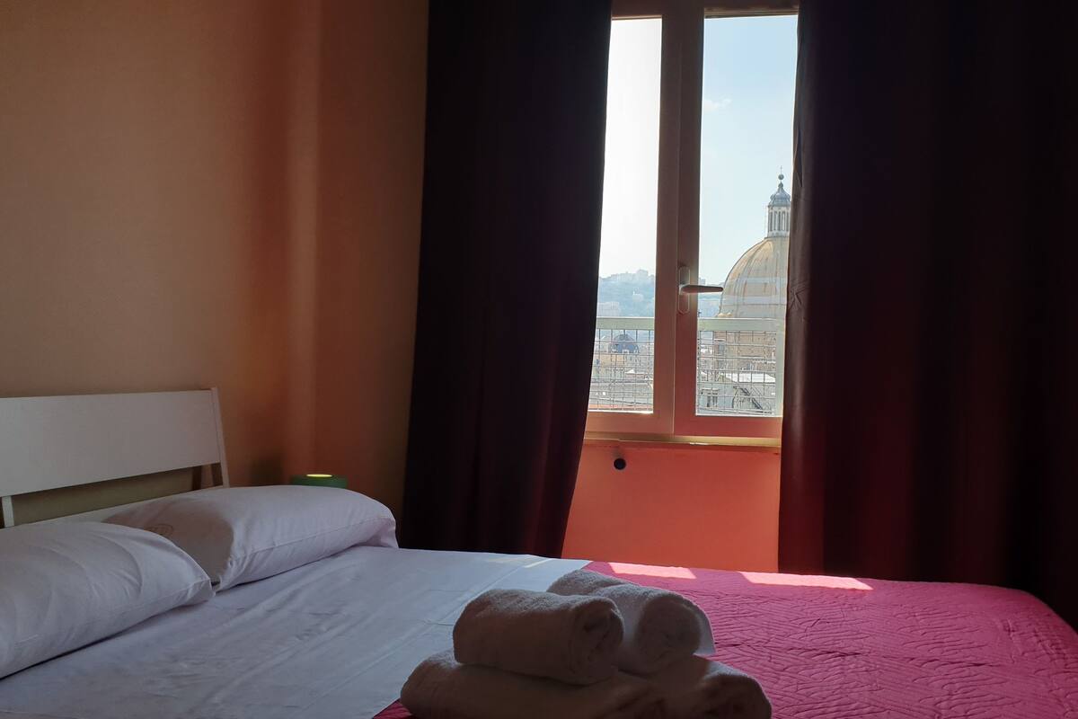A hotel room with a bed, pillows, a window with curtains, and a view of a building with a dome.