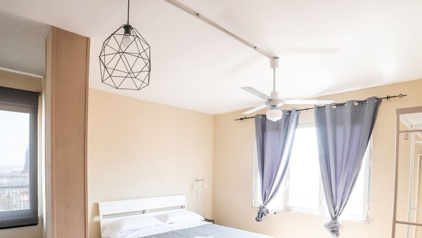 Une chambre à coucher avec un lit, un ventilateur de plafond, une fenêtre avec des rideaux et une vue sur la ville.