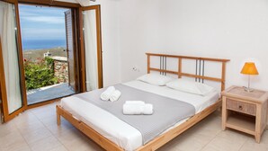 A bedroom with a wooden bed, white bedding, a nightstand with a lamp, and a view of the sea through an open door. - Karthea VI (Kea)