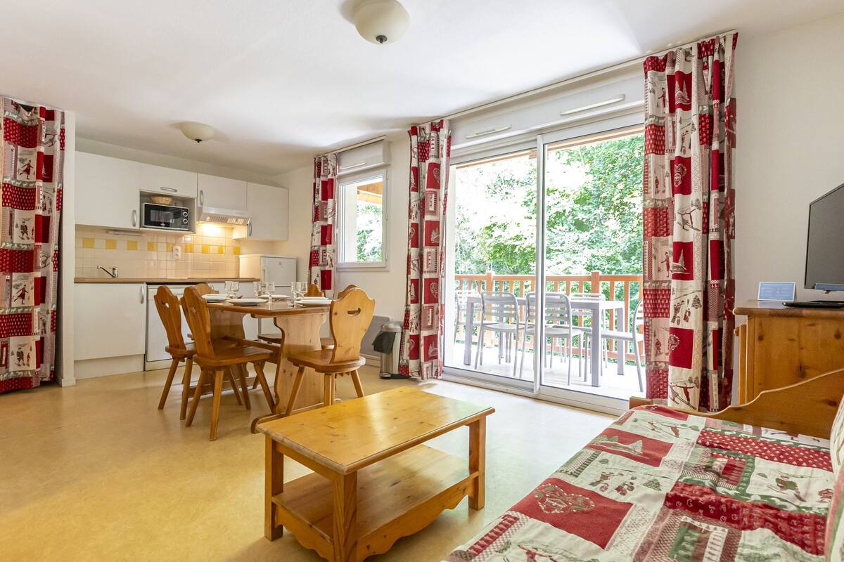 A cozy room with a wooden table and chairs, a bed with a quilt, a TV, and a balcony with chairs.