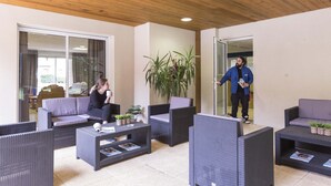 A covered outdoor seating area with wicker furniture, a potted plant, and a glass door leading to an interior space. - Résidence Odalys Soleil d'Aure (Saint-Lary-Soulan)