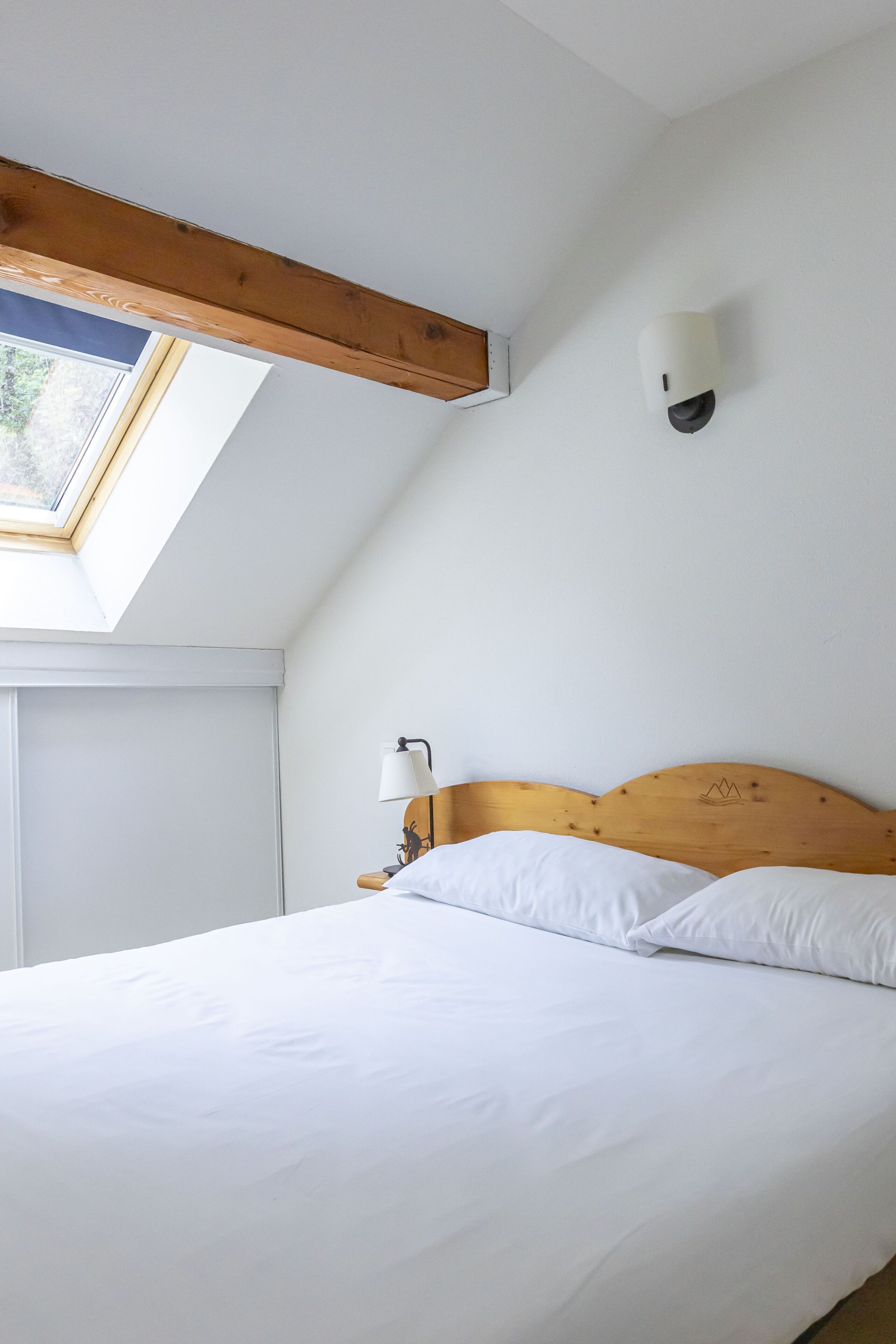 A bedroom with a wooden headboard, a skylight, a bedside lamp, and white bedding.
