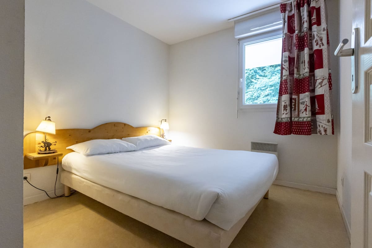 A neatly made bed in a room with a wooden headboard, a nightstand with a lamp, a window with a view of greenery, and a red and white patterned curtain.