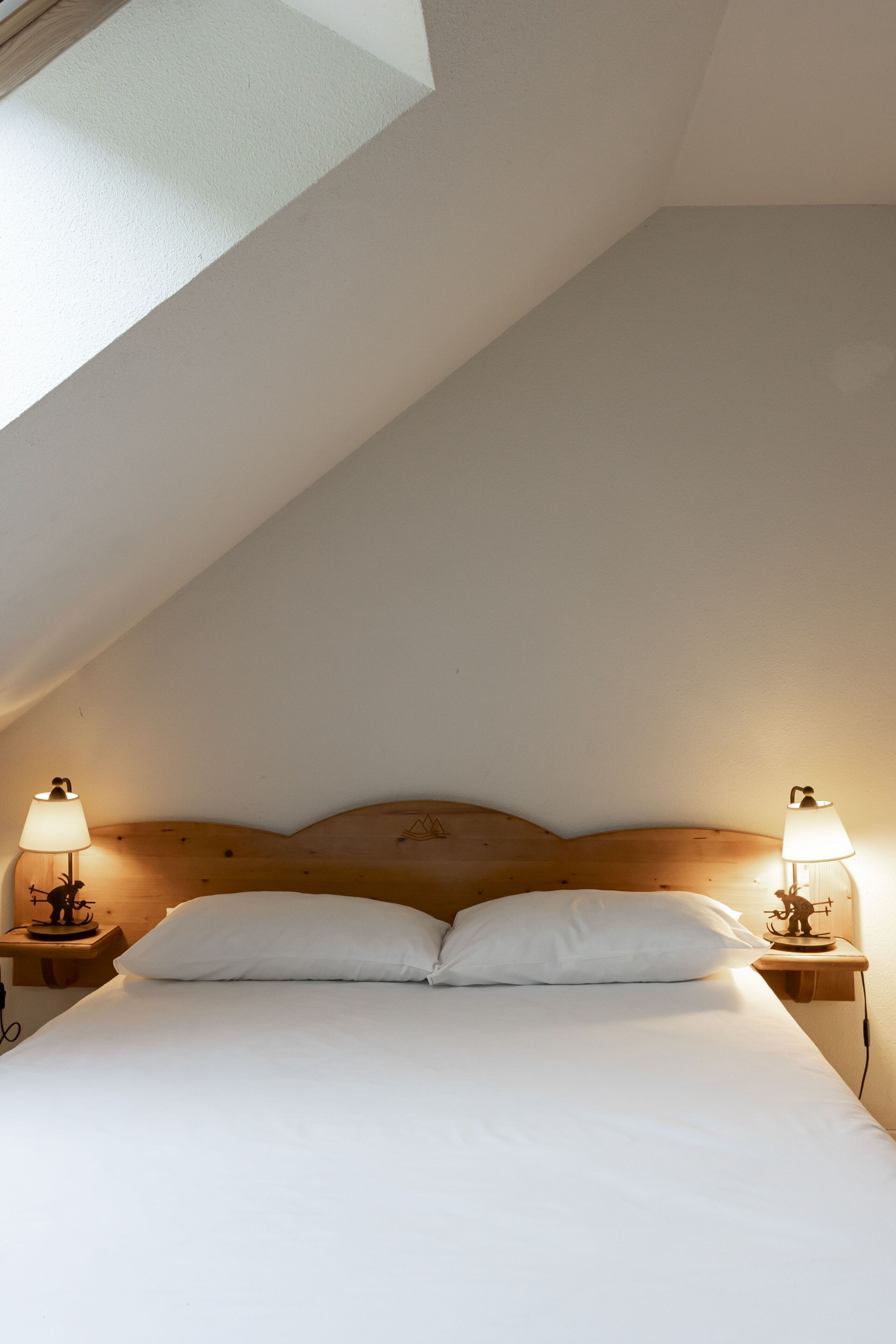 Un dormitorio con techo inclinado, cabecera de madera, dos lámparas de noche y cubrecama blanco.