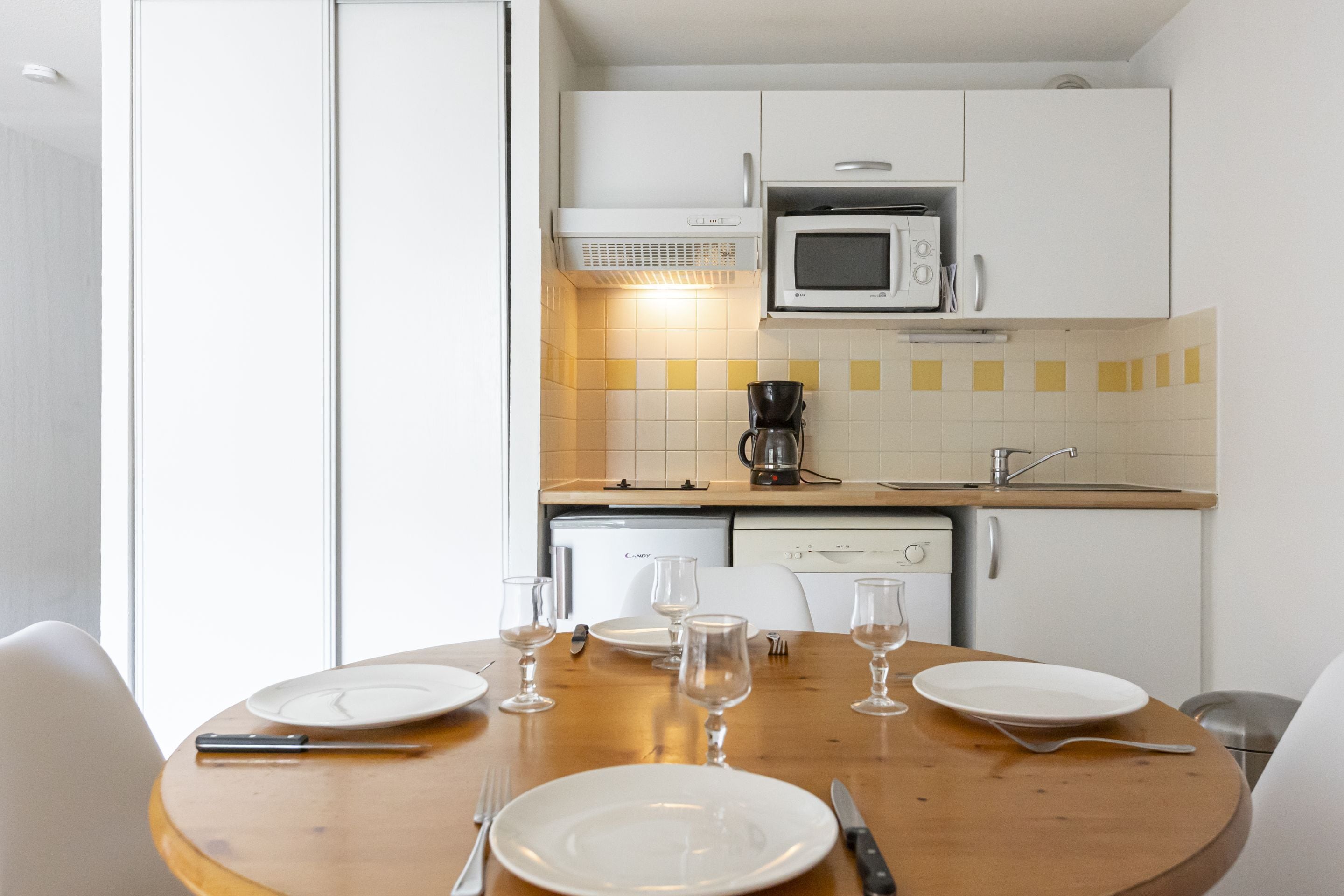 Uma mesa de jantar posta com pratos, talheres e copos de vinho em uma cozinha com armários brancos, micro-ondas e cafeteira.