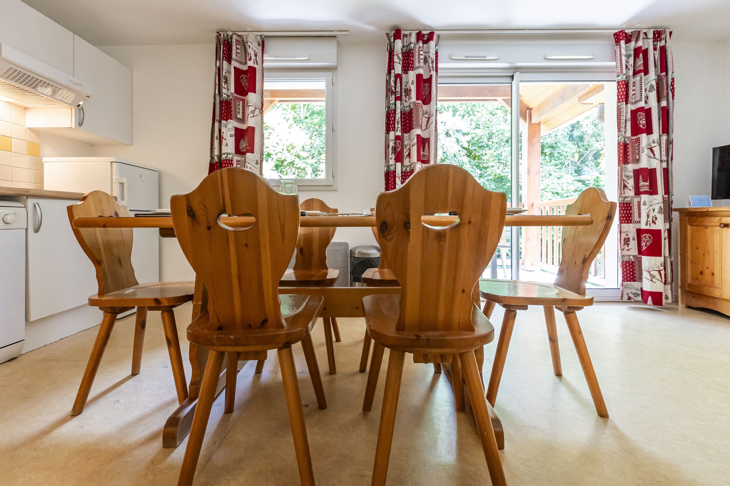A dining area with wooden chairs and a table, white cabinets, and a view through a window with patterned curtains.