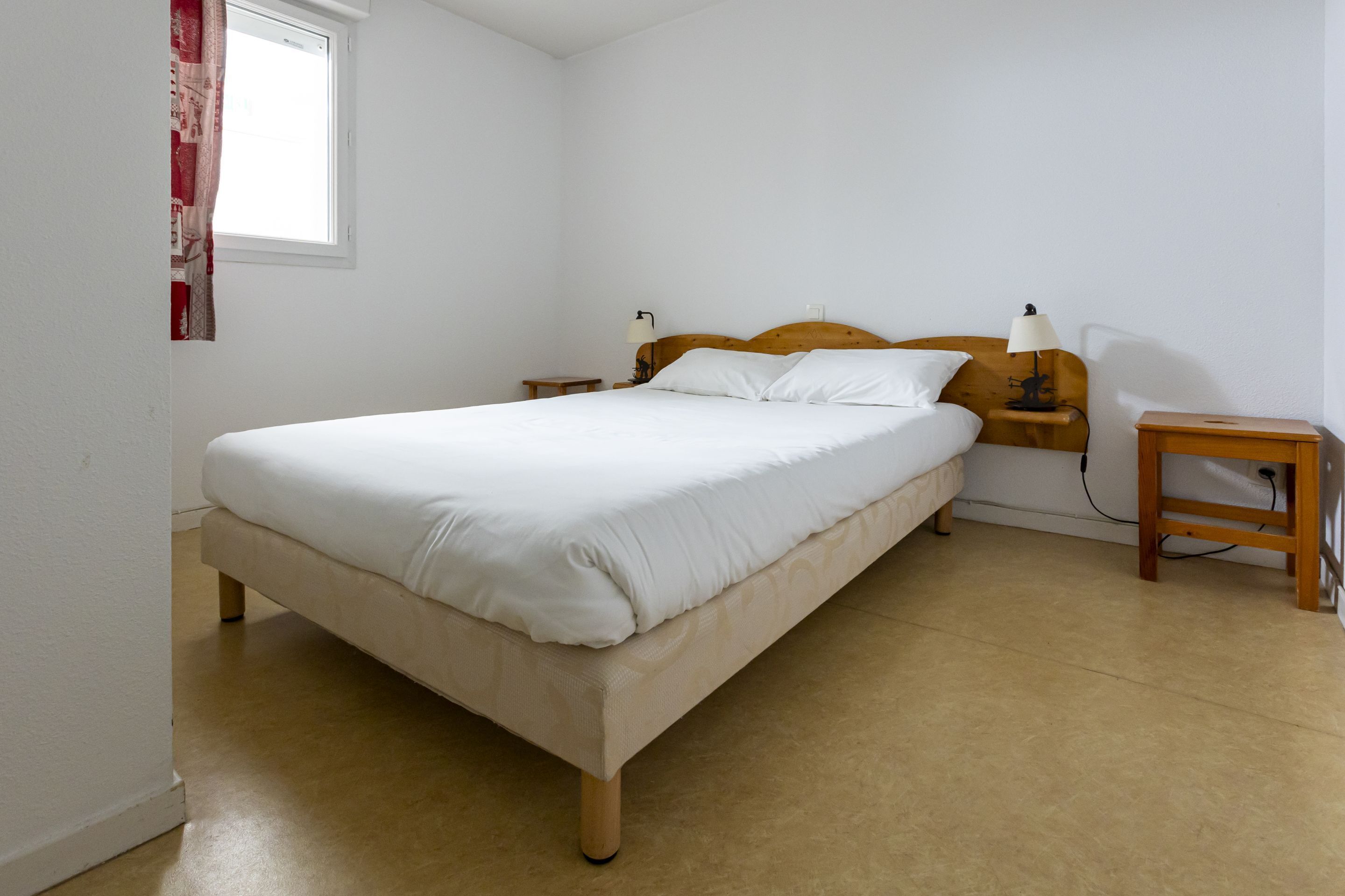 Une chambre à coucher comprenant un lit, une tête de lit en bois, deux tables de chevet avec des lampes, une petite table de nuit en bois et une fenêtre avec des rideaux.