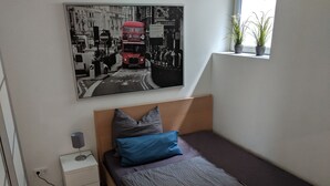 A small bedroom with a single bed, a nightstand, a lamp, and a wall-mounted picture of a city street scene. - Business Apartments Ratingen 5 (Ratingen)