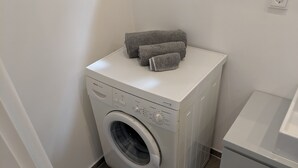 A compact, white Bosch washing machine in a small laundry room with a tiled floor and a white cabinet. - Business Apartments Ratingen 5 (Ratingen)