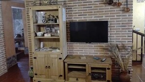 A living room with a brick wall, a wooden cabinet with shelves, a flat-screen TV mounted on the wall, and a small table with a vase of dried plants.