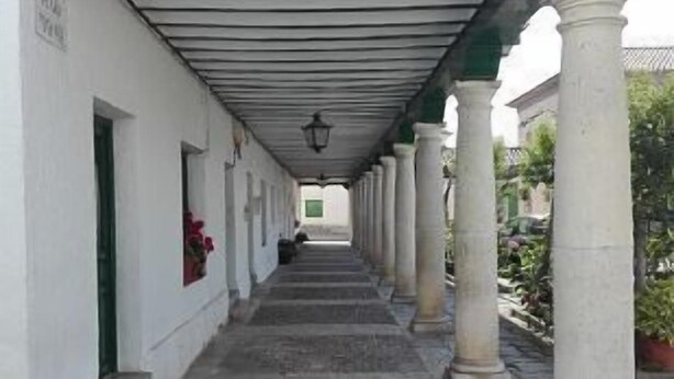 A covered corridor with stone flooring, white walls, and green trim. Columns line the corridor, and there is a sign on the wall.