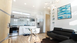 Una cocina moderna con electrodomésticos de acero inoxidable, una zona de comedor con mesa y sillas de vidrio, y un sofá oscuro en un espacio habitable luminoso.