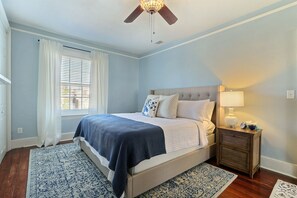Une chambre à coucher avec un lit, une table de chevet, un ventilateur de plafond et une fenêtre avec des rideaux.