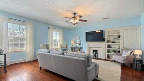A living room with a grey sofa, wooden flooring, a ceiling fan, a fireplace, and a TV mounted on the wall. - Private Parking and Unbeatable Location on Liberty Street by Lucky Savannah (Savannah)