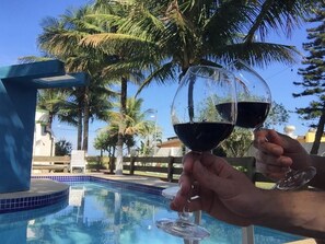 A poolside view with a clear sky, palm trees, and a person holding a wine glass. - Pousada Icaraí - Ilha Comprida (Ilha Comprida)