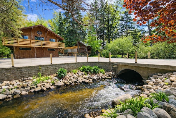 Une cabane en bois avec un pont de pierre enjambant un ruisseau, entourée d’arbres et de verdure.