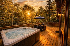 A hot tub on a wooden deck with a table and chairs, surrounded by trees and a sunset view. - WalkToBeach HotTub Fireplace Authentically North (Traverse City)