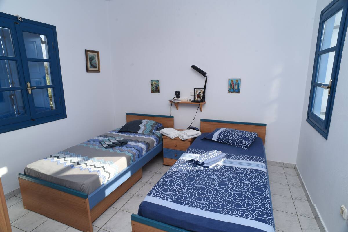 Une chambre avec deux lits simples, une table de chevet, une étagère murale avec des livres et une lampe, et de petites photos encadrées accrochées au mur.