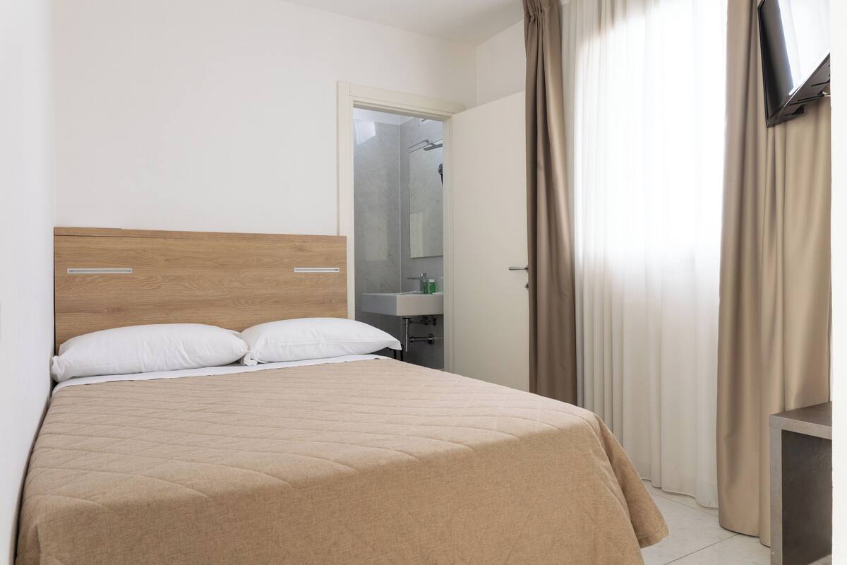 A hotel room with a bed, a wooden headboard, a bedside table, a mirror, and a bathroom visible through an open door.