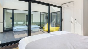 Ein modernes Hotelzimmer mit einer großen Schiebeglasdoor zum Balkon, einem Bett mit weißer Bettwäsche und einem gelben Kopfkissen.