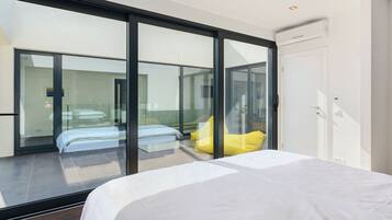 A modern hotel room with a large sliding glass door leading to a balcony, a bed with white bedding, and a yellow pillow.