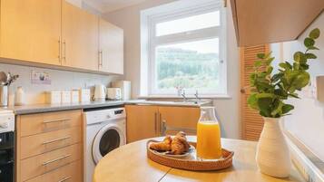 Une cuisine avec des meubles en bois, un lave-linge, une fenêtre donnant sur un espace verdoyant, une table avec des croissants et du jus d’orange, et une plante en pot.