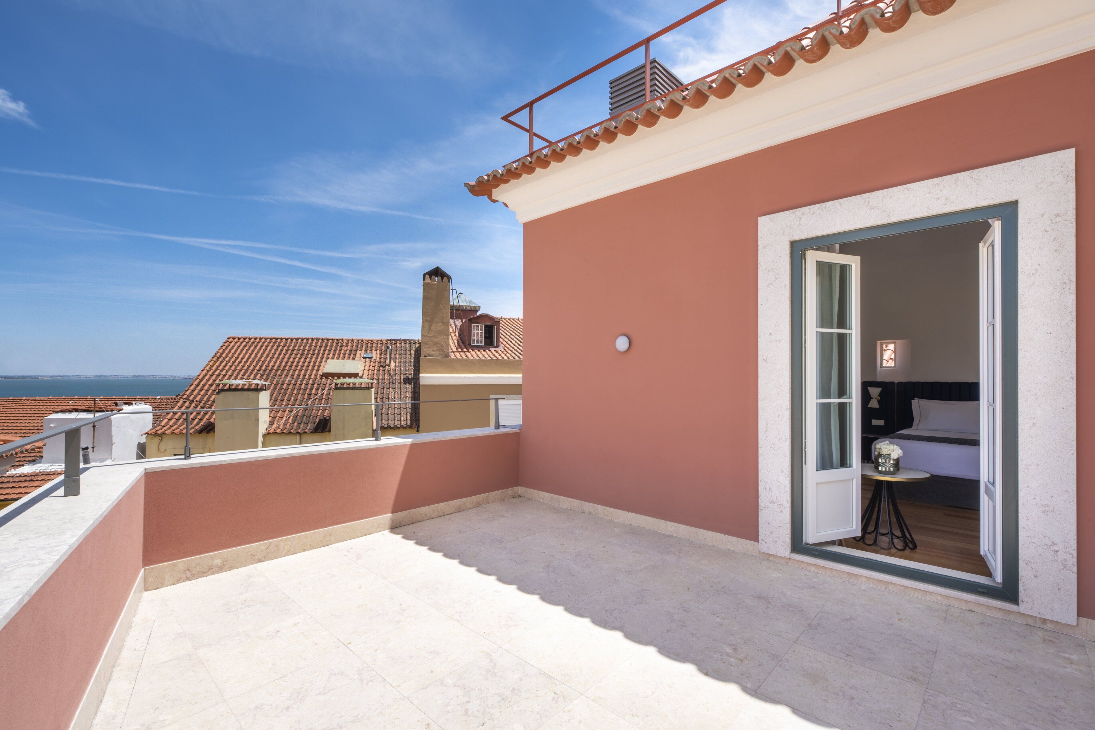 Una terraza en la azotea con vistas despejadas al mar, un balcón con barandilla y una puerta que da a una habitación con cama y una mesita.