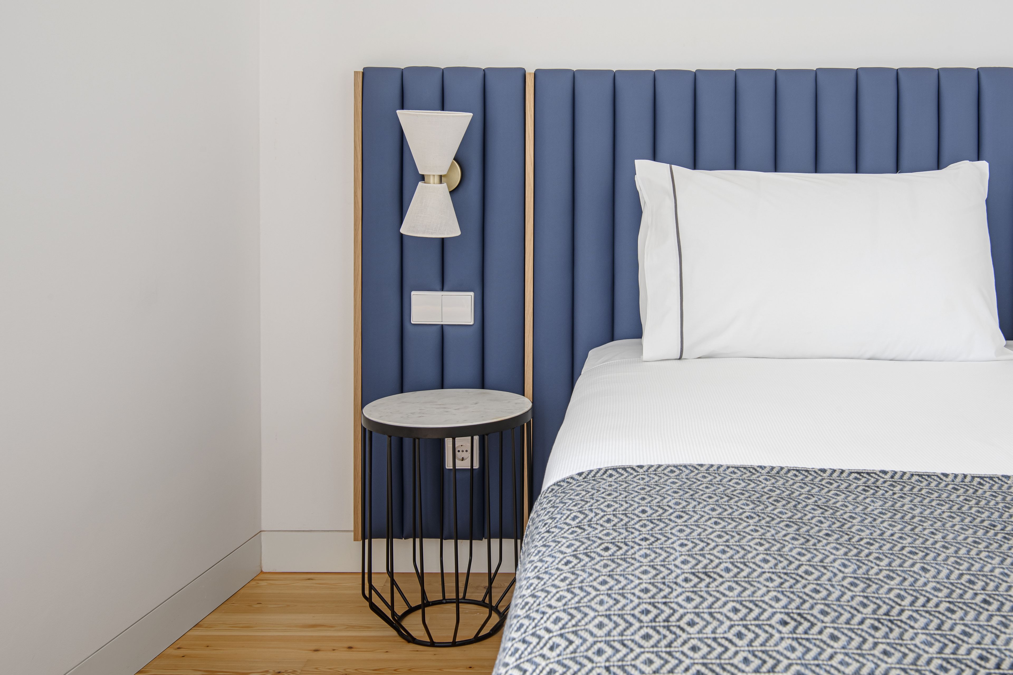 Un dormitorio con cabecera azul, una mesita redonda, una lámpara blanca y una colcha estampada.
