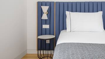 Un dormitorio con cabecera azul, una mesita redonda, una lámpara blanca y una colcha estampada.