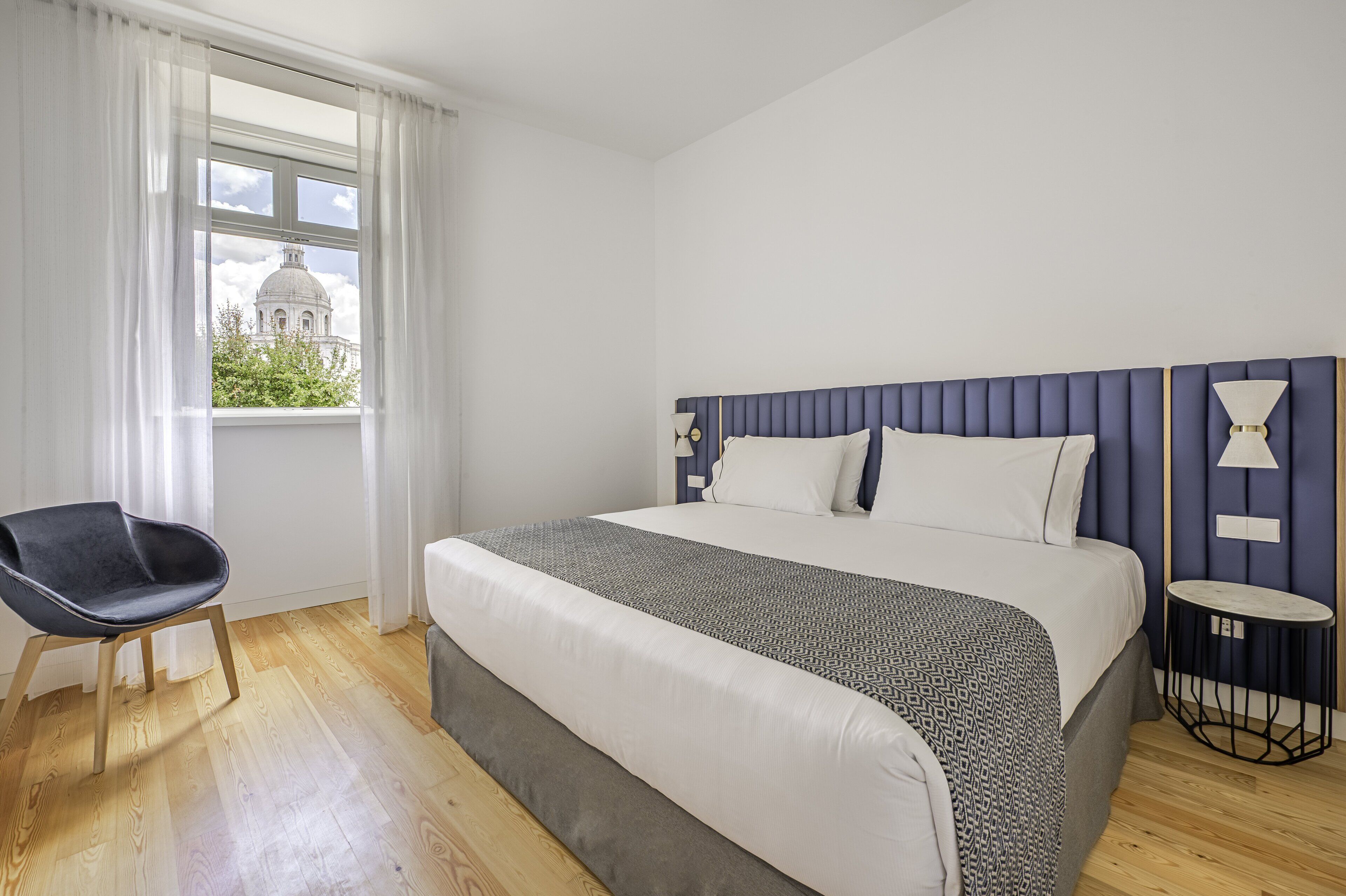 Une chambre à coucher avec un grand lit, une chaise, une table de chevet et une fenêtre donnant sur un immeuble.