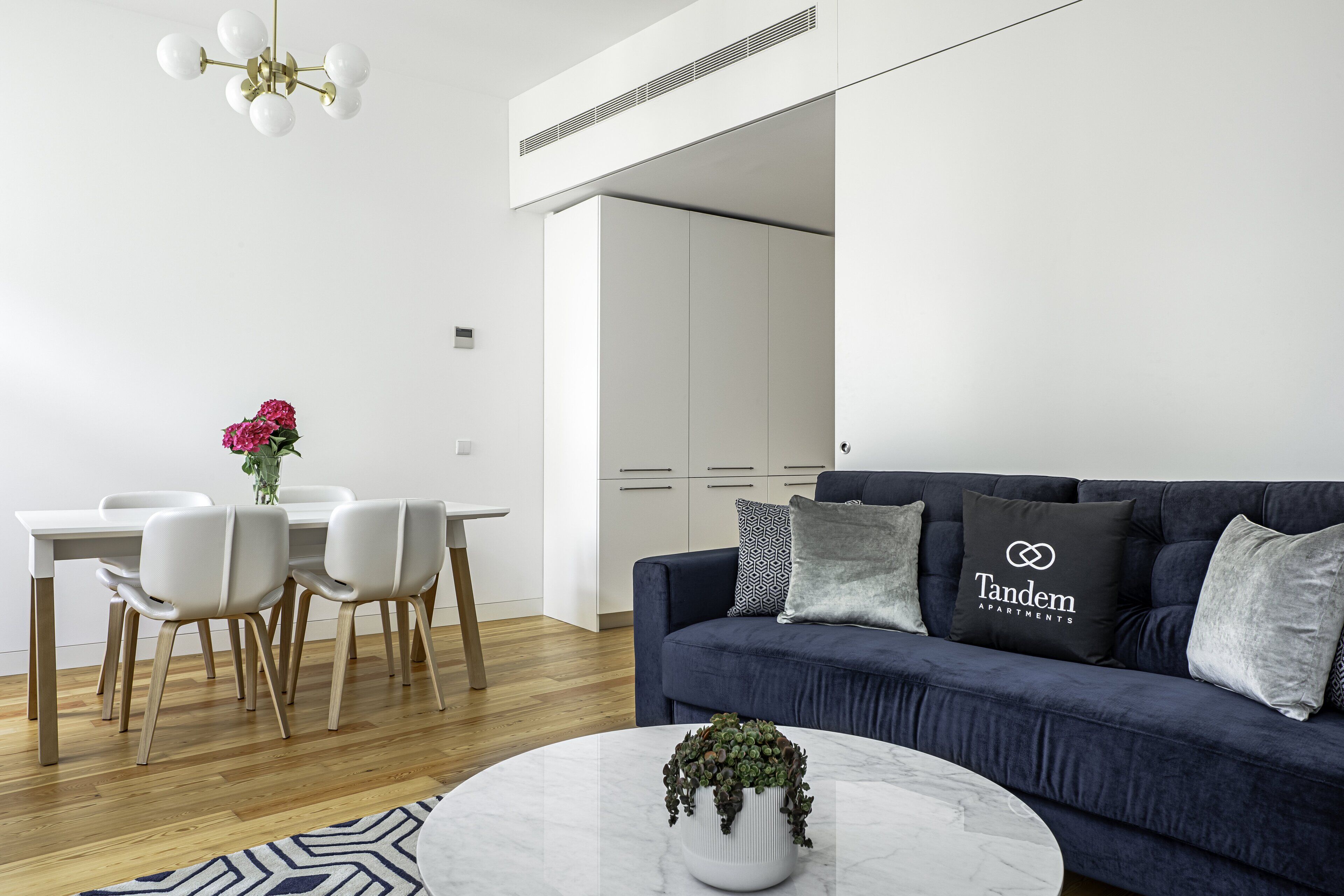 A modern living room with a dark blue sofa, a white dining table with chairs, and a marble coffee table with a plant.