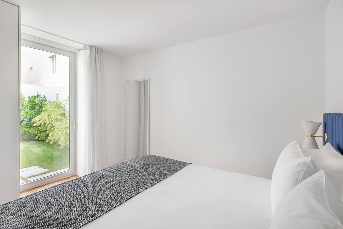 Un dormitorio moderno con una cama grande, vistas a un jardín a través de una puerta corredera y una mesita de noche con lámpara.