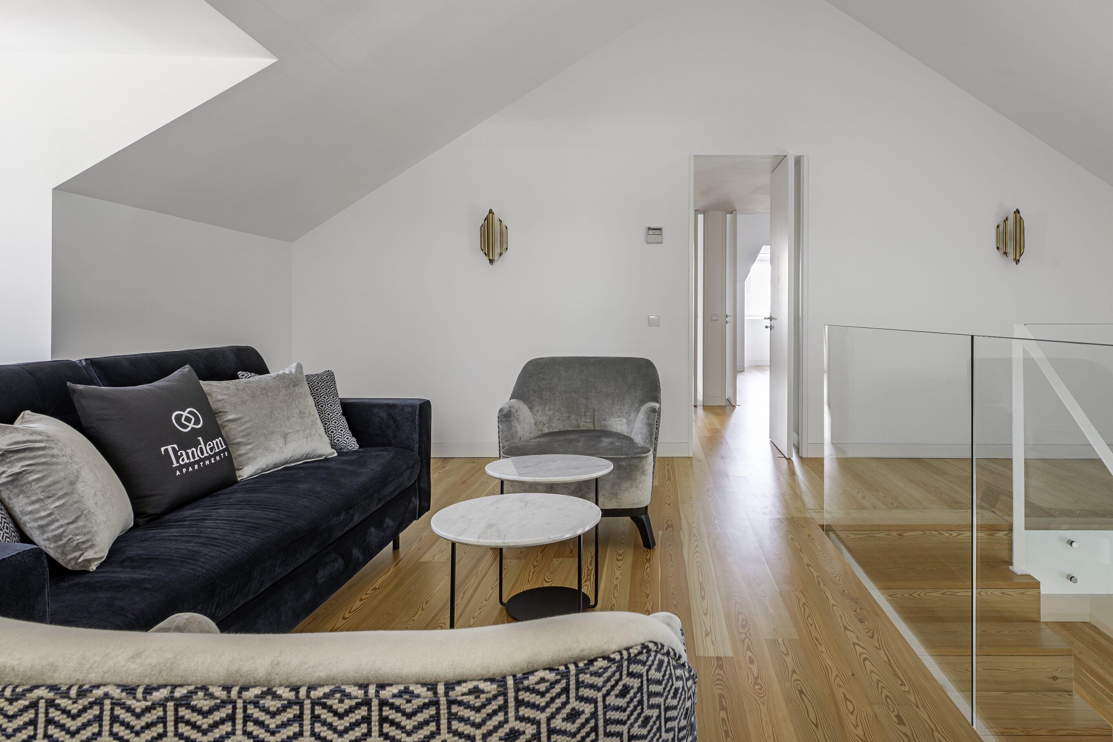 A modern living room with a dark sofa, a grey armchair, and a small round table. A staircase with a glass railing leads to an upper floor.