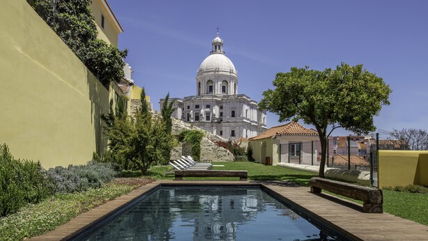 Una piscina con cubierta de madera, rodeada de vegetación y un edificio con cúpula al fondo.