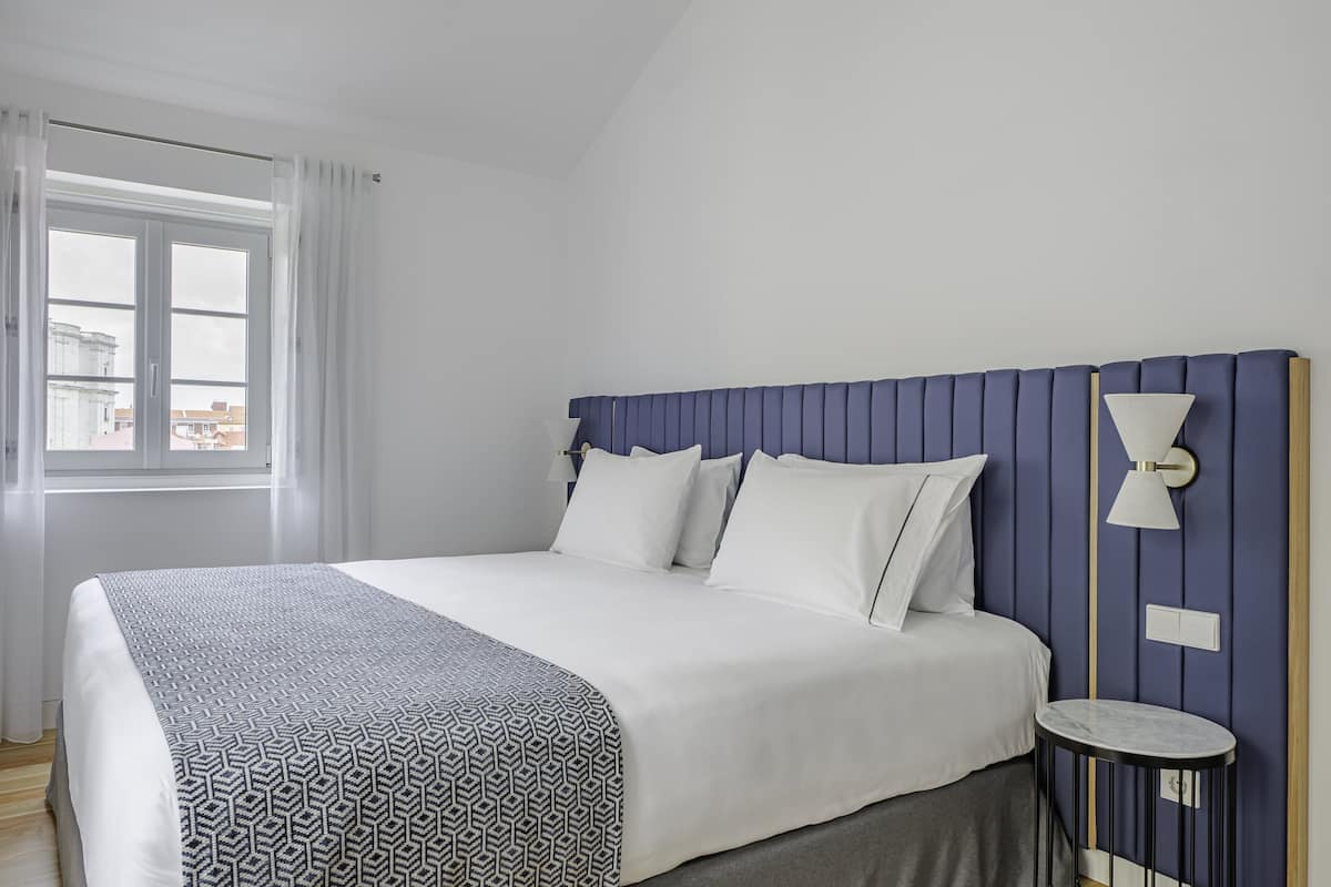 Una cama bien hecha con un edredón estampado, una lámpara de noche y vistas de edificios a través de una ventana.