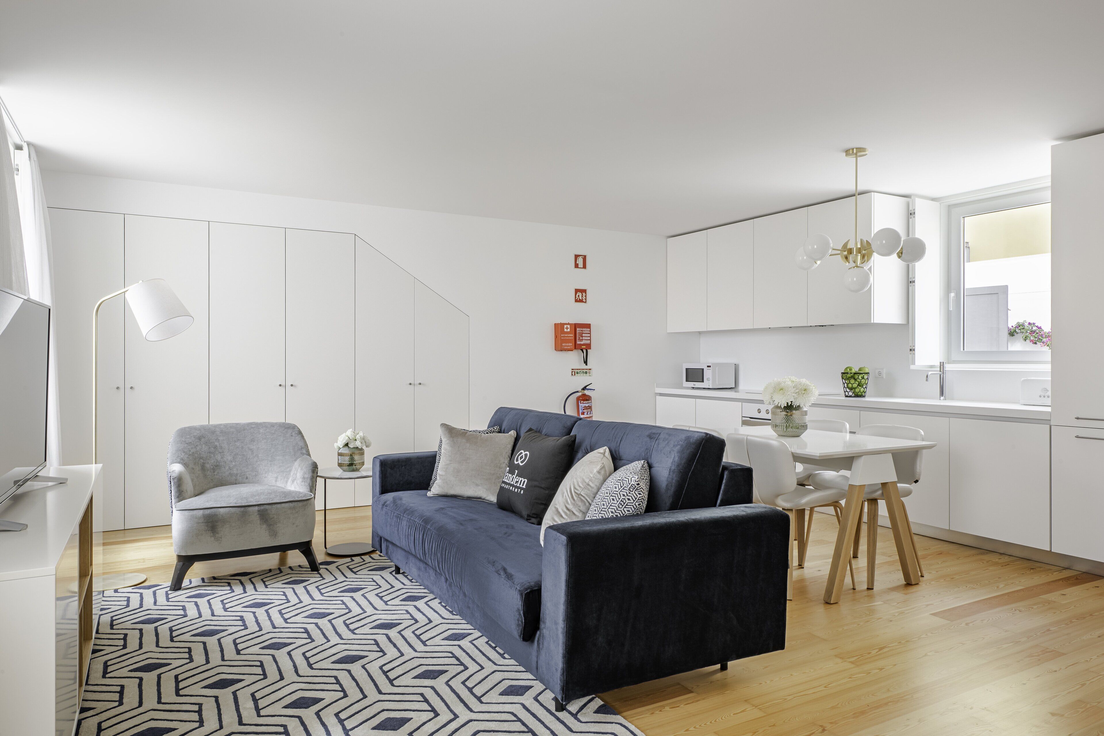 A modern living room with a blue sofa, a grey armchair, and a white dining table. There is a patterned rug, a small side table, and a pendant light. The room has white walls and a wooden floor.