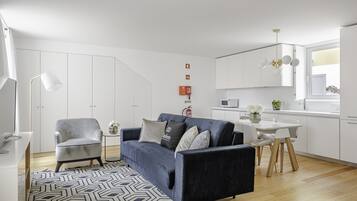 A modern living room with a blue sofa, a grey armchair, and a white dining table. There is a patterned rug, a small side table, and a pendant light. The room has white walls and a wooden floor.