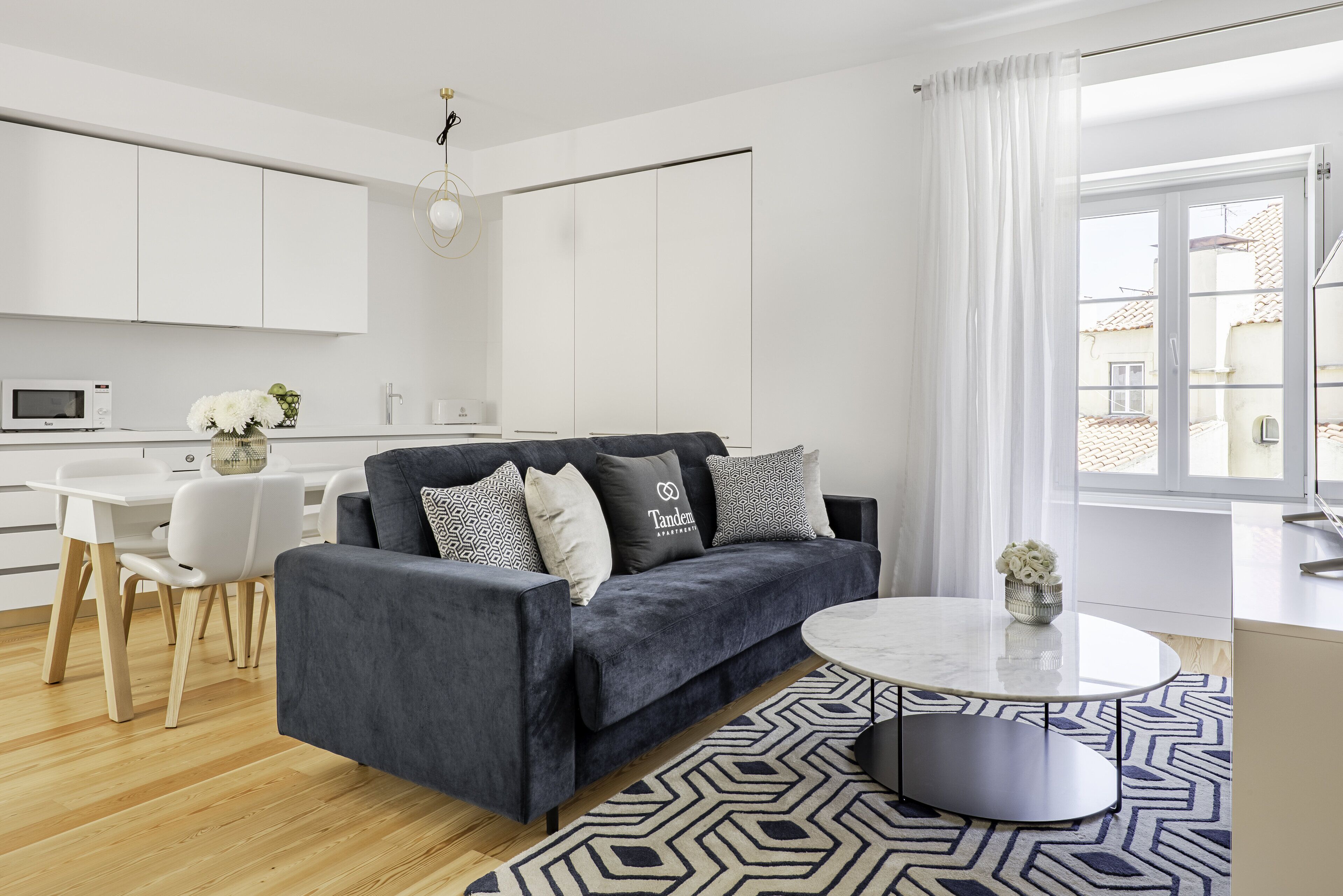 A modern living room with a dark grey sofa, white dining table, and a patterned rug.