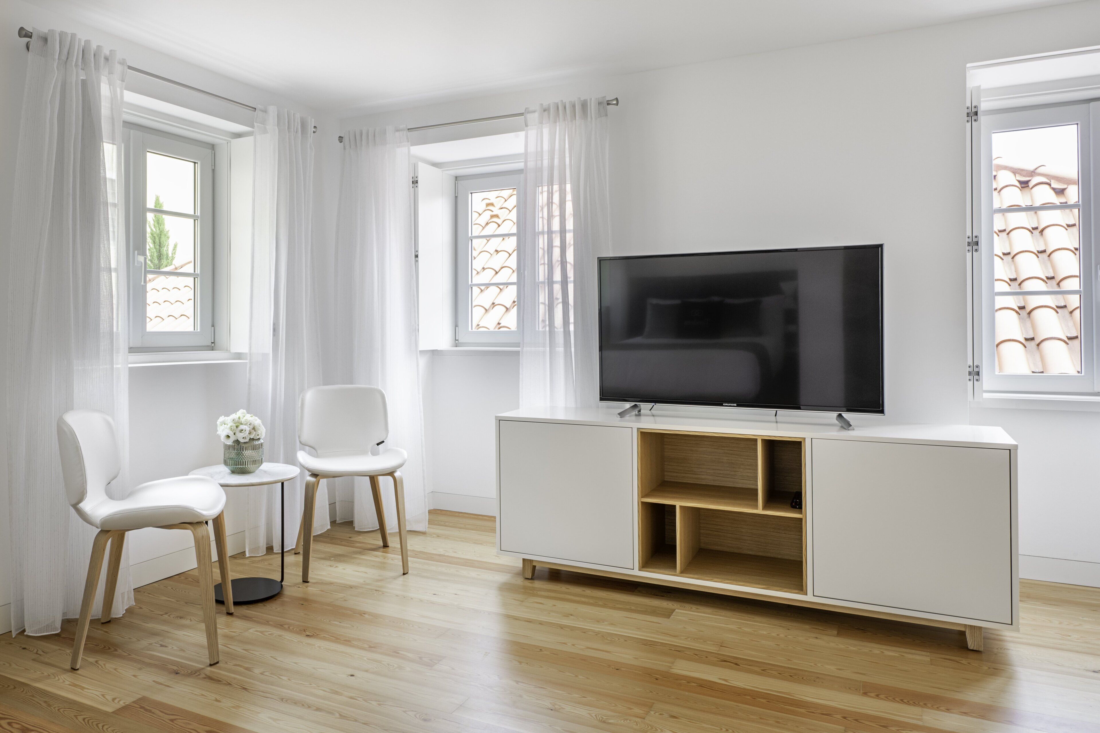 A modern living room with a flat-screen TV on a white cabinet, two white chairs, a small round table with a vase, and large windows with white curtains.