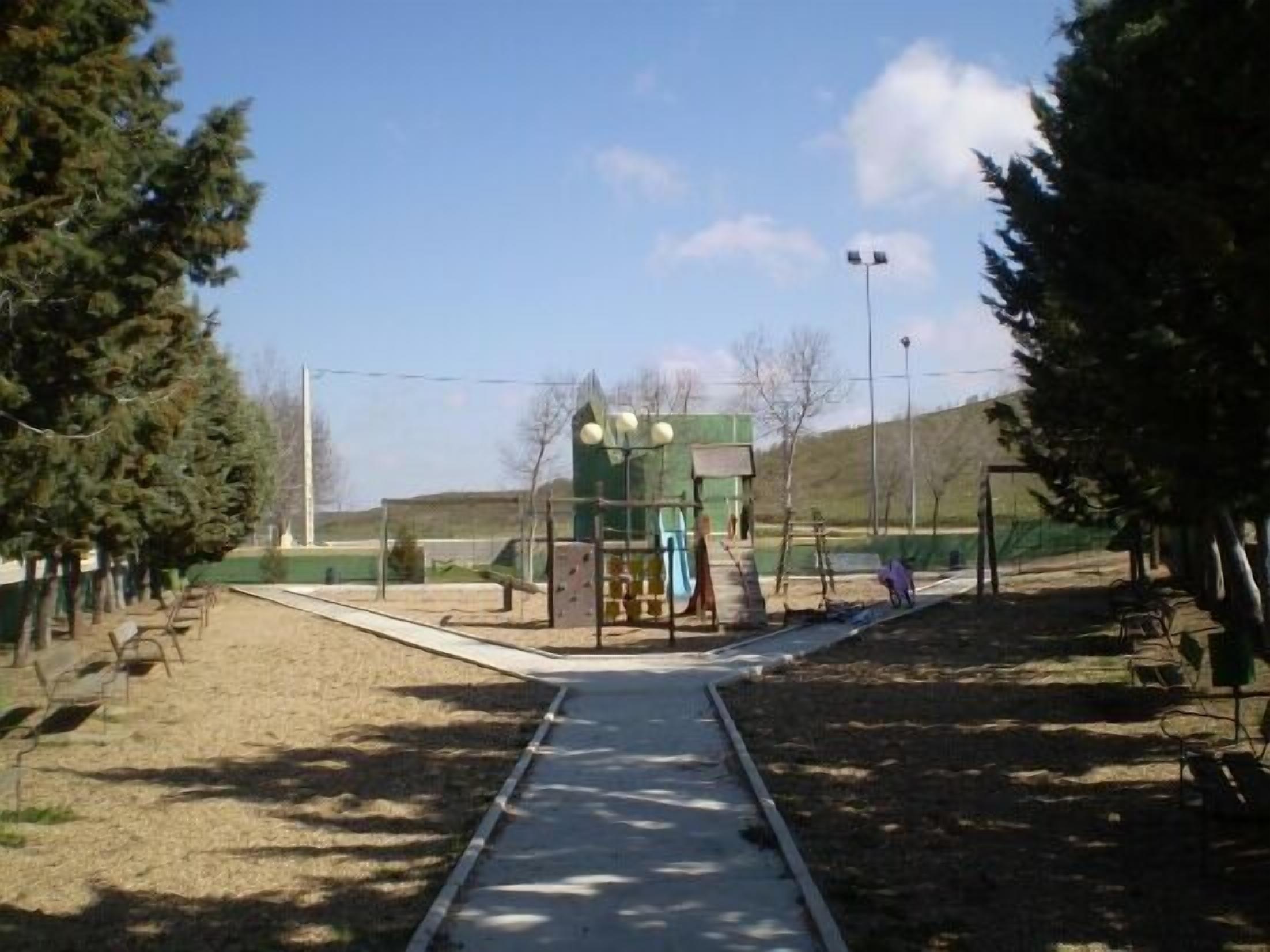 A pathway leading to a playground with a green building, surrounded by trees and a clear sky.