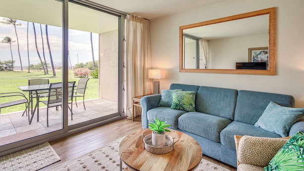A living room with a sofa, a coffee table with a plant, a mirror, and a view of a grassy outdoor area through large windows. - Gorgeous Oceanfront Condo with Spectacular Views! (Kihei)