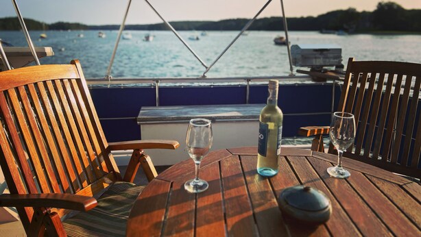 A wooden table with two wine glasses and a bottle, set under a canopy on a boat with a view of the water and boats in the distance. - Maine Bed & Boat, MAZU on the scenic Damariscotta River (Damariscotta)