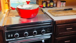 A kitchen with a red pot on a stove, a wooden cabinet, and a window with a view of a lighthouse. - Maine Bed & Boat, MAZU on the scenic Damariscotta River (Damariscotta)