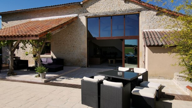 A stone building with a tiled roof, a large glass door, outdoor seating, and a pergola with plants. - Restored barn from late 18th century with pool (Champniers)