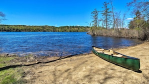 Una canoa en la orilla arenosa de un lago tranquilo con árboles al fondo.