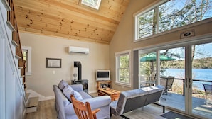 Una sala de estar acogedora con un sofá, sillas de madera, un televisor y una chimenea. Amplias ventanas ofrecen vistas al lago.