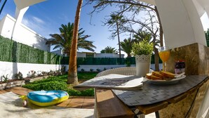 Un patio con una mesa dispuesta para el desayuno, que incluye tostadas y un vaso de zumo de naranja, rodeado de follaje y palmeras.