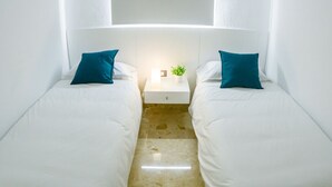 Habitación de hotel con dos camas individuales, ropa de cama blanca, almohadas verde azulado y una mesita de noche pequeña con una planta, además de un cabecero blanco.