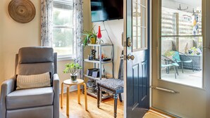 A living room with a grey leather chair, a small table with a plant, a bookshelf, and a TV on the wall. - Nags Head Apt with Patio - 1/2 Mile Walk to Beach! (Nags Head)