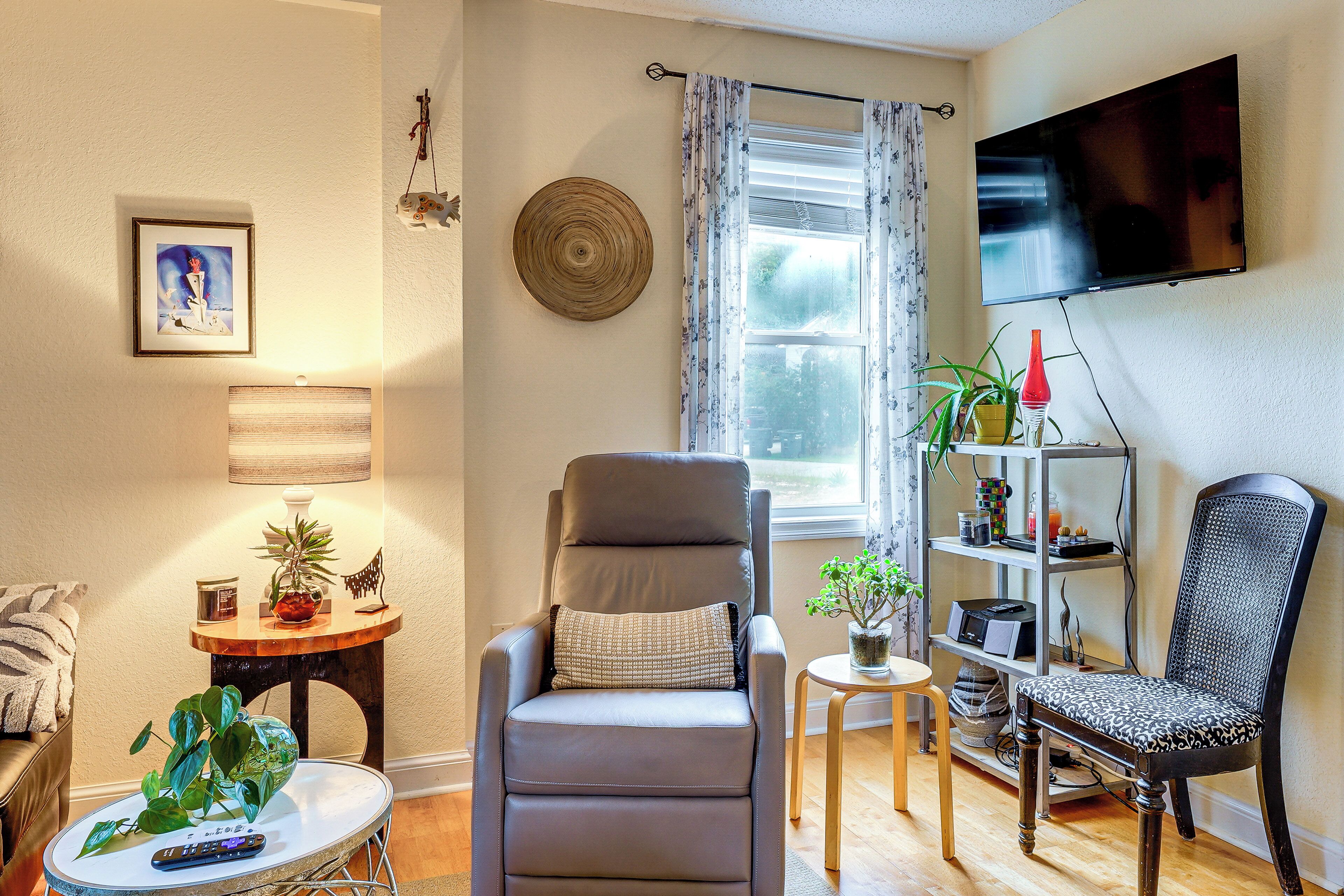 A living room with a grey armchair, a small round table, a bookshelf, a flat-screen TV, and a window with curtains.